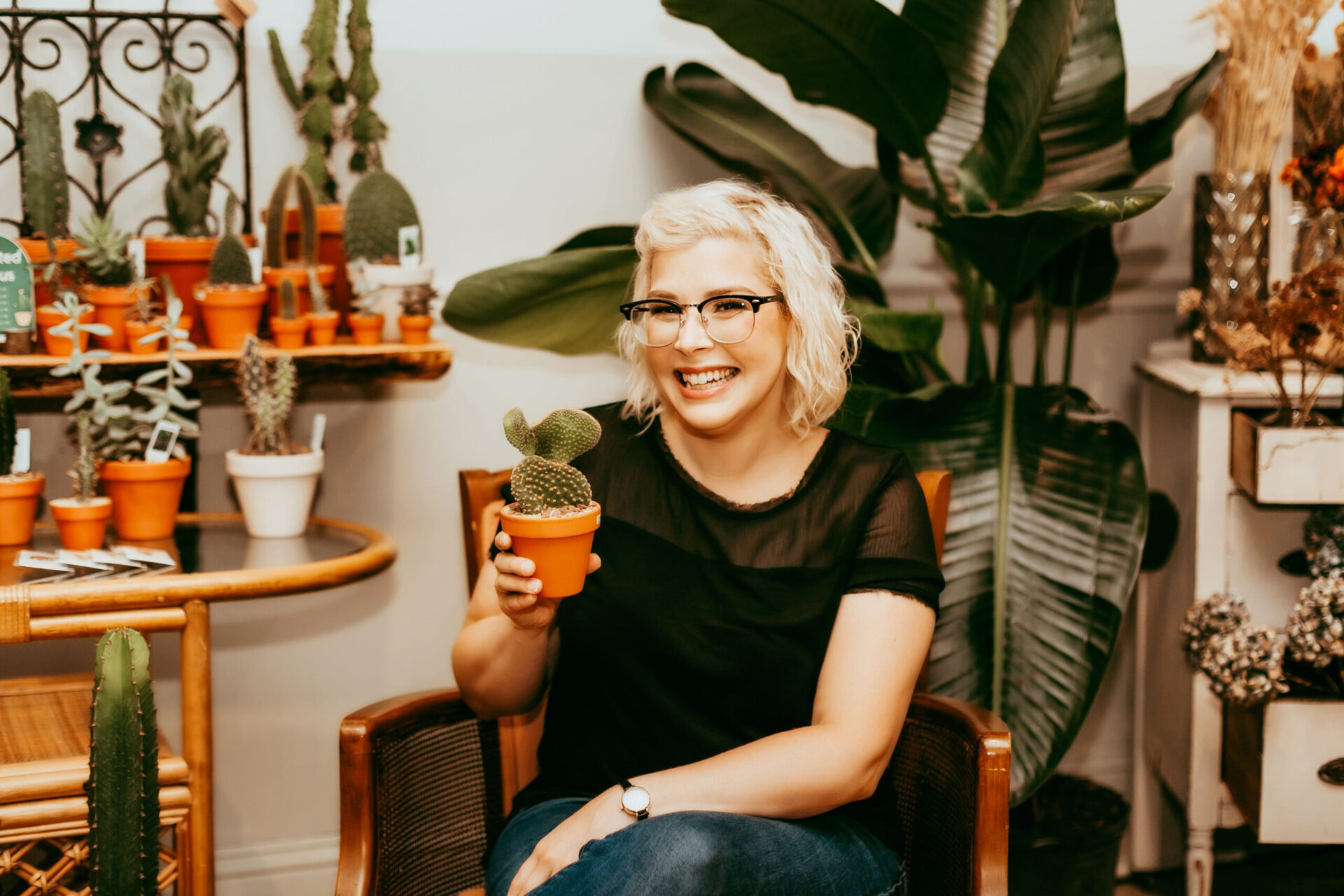Christina, a pale white woman wearing a black top, jeans, and browline glasses sits in an armchair with wooden armrests. She is surrounded by plants, and is holding a small potted cactus and smiling.