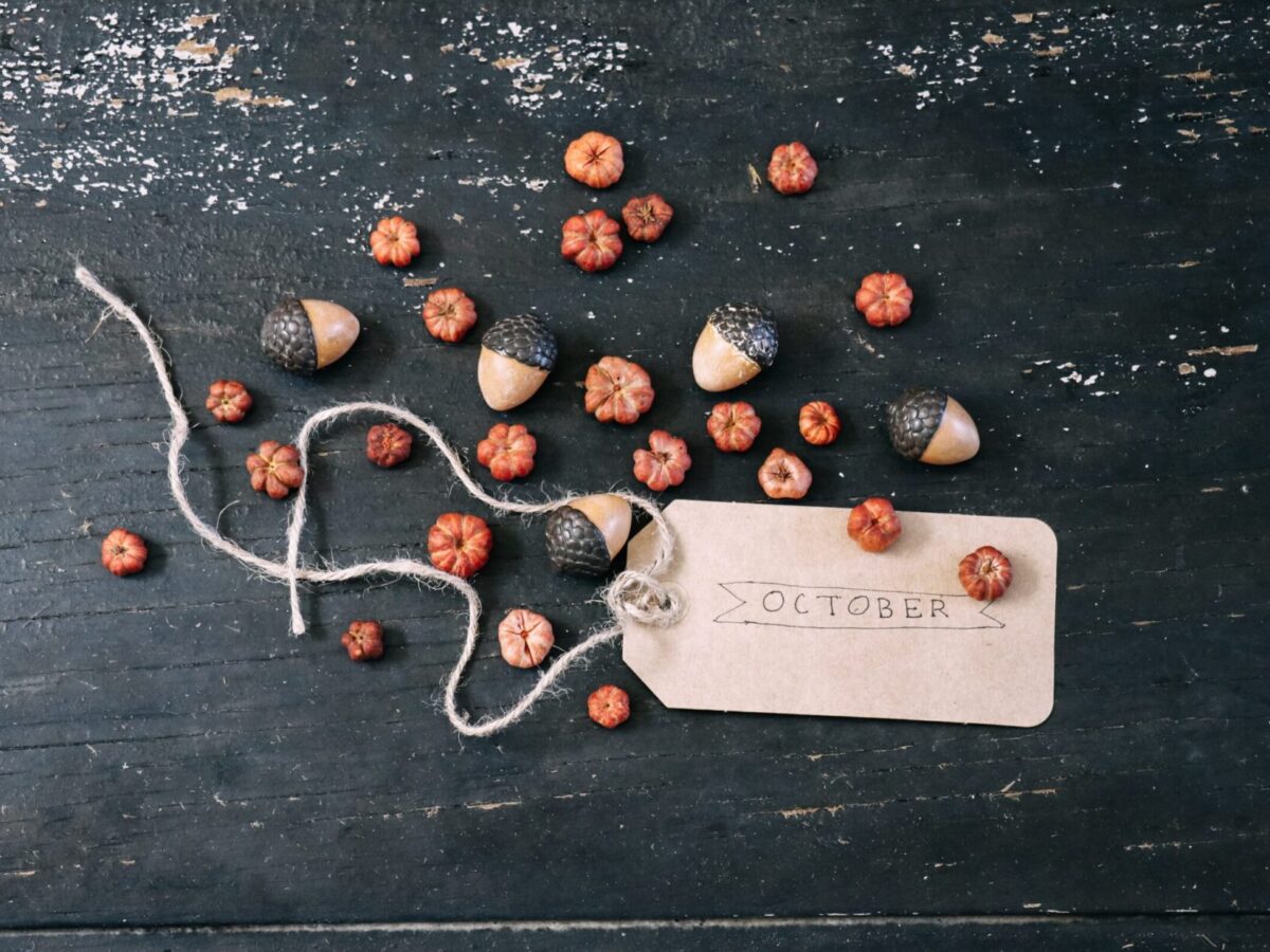 A handful of tiny pumpkins and acorns are scattered on a distressed black table, surrounding a paper tag reading "October."