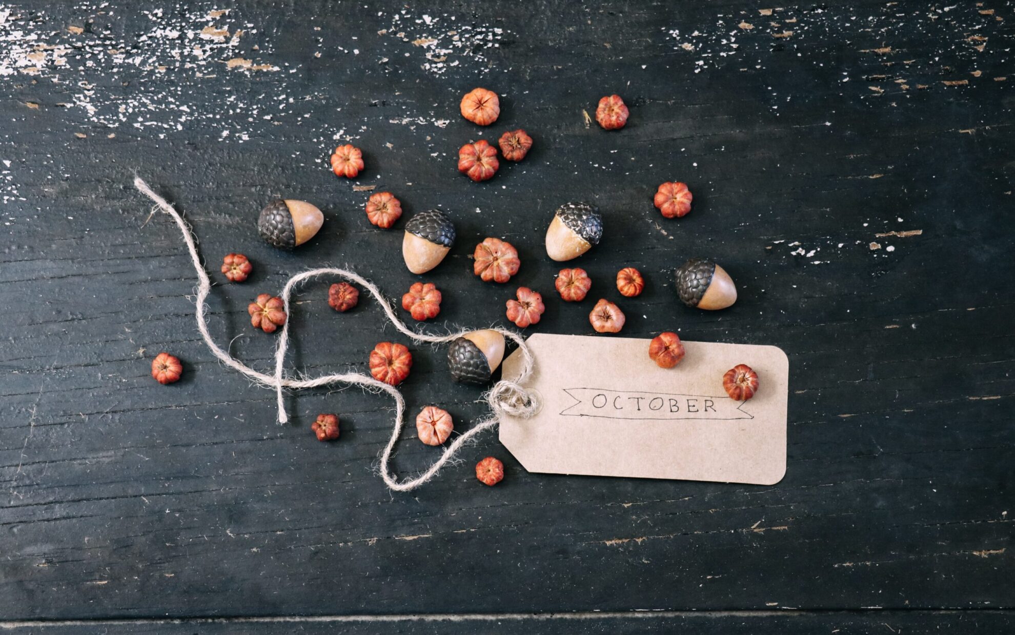 A handful of tiny pumpkins and acorns are scattered on a distressed black table, surrounding a paper tag reading "October."