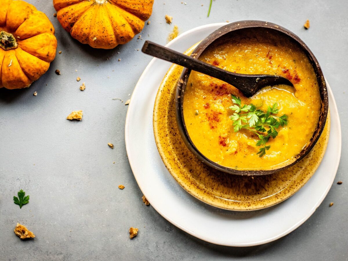 A generous bowl of orange soup, speckled with greek herbs and red seasoning, rests atop two plates, one yellow and the larger one white. In the upper left corner are two smaller pumpkins.