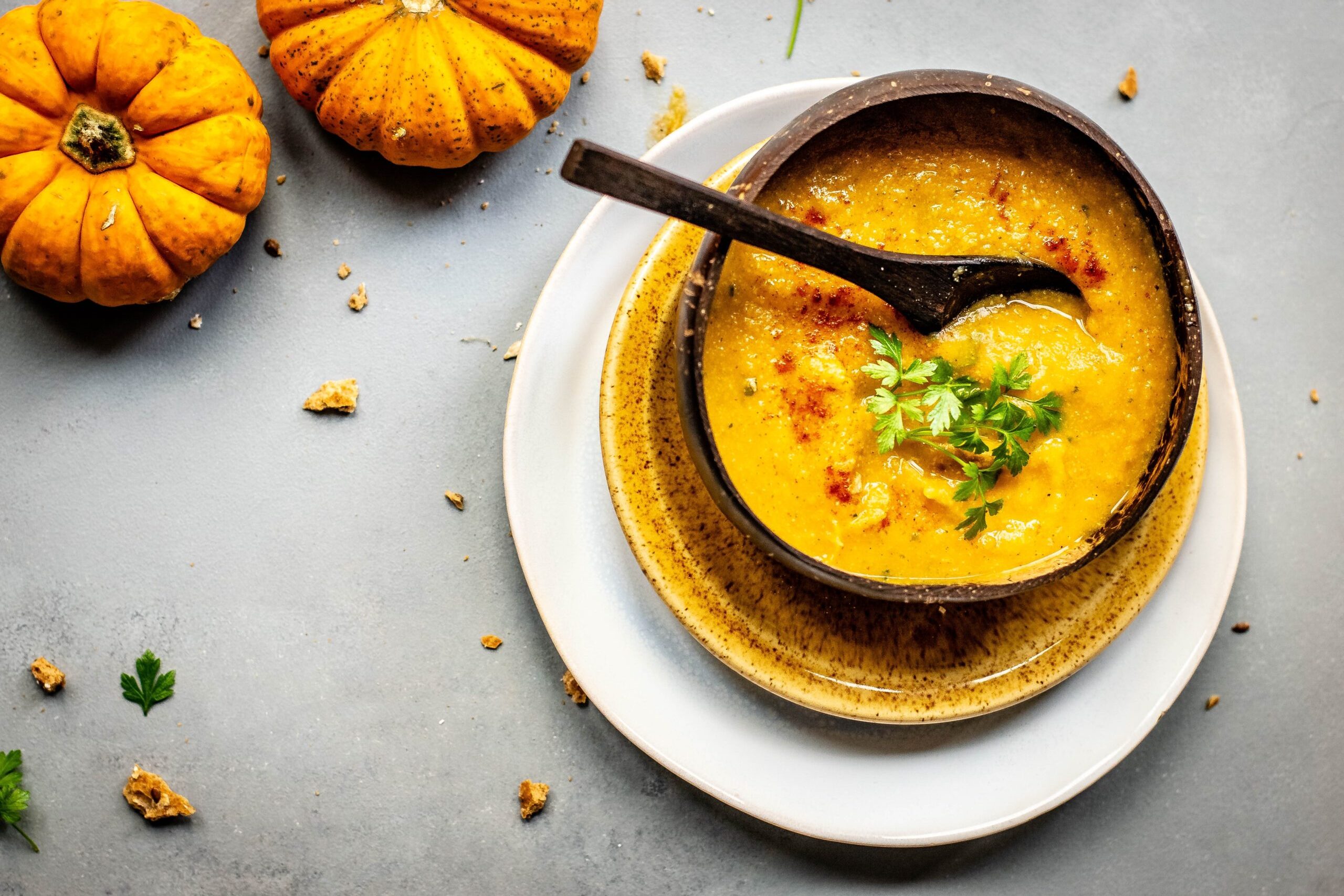 A generous bowl of orange soup, speckled with greek herbs and red seasoning, rests atop two plates, one yellow and the larger one white. In the upper left corner are two smaller pumpkins.