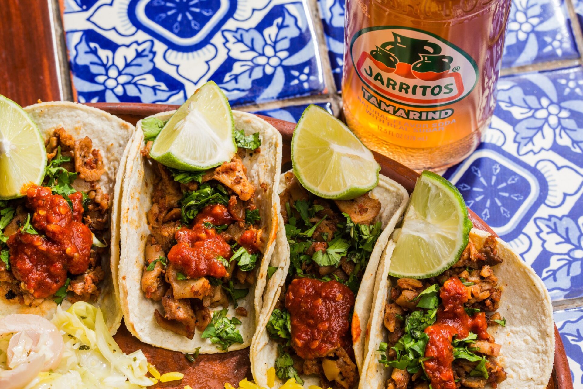 A plate of four tacos with lime slices sits on a table next to an orange Mexican soda.