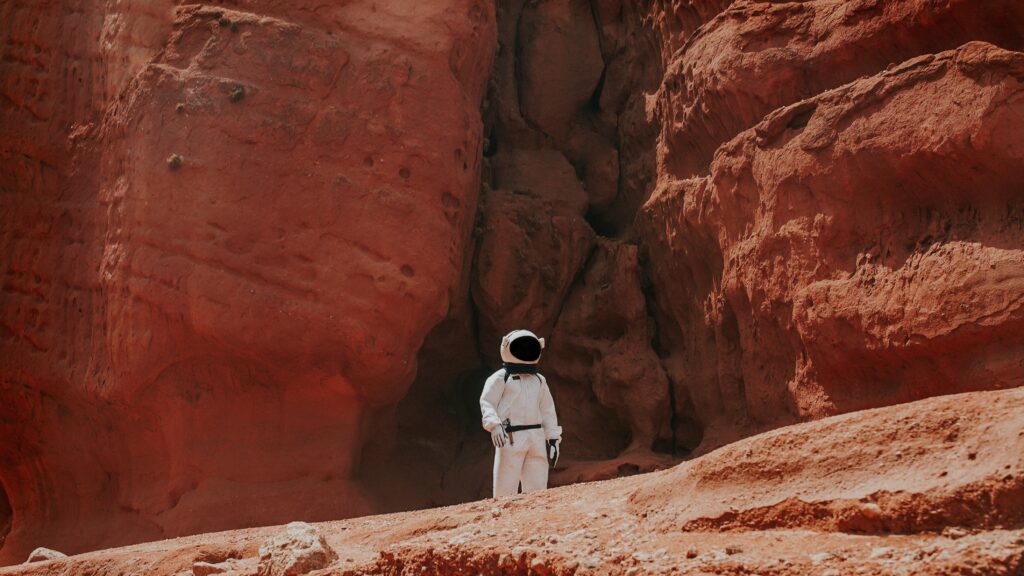 An astronaut in a white space suit wanders a red, dusty, rocky terrain.