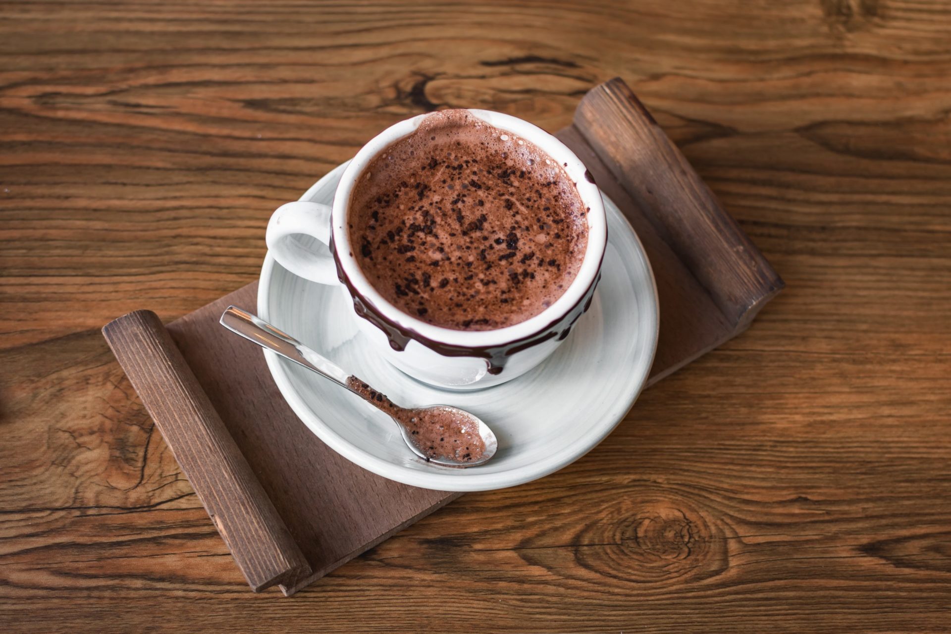 A cup of hot chocolate in a white mug rimmed with chocolate sits atop a white plate, wooden serving tray, and wooden table. There is a chocolate covered spoon on the plate.