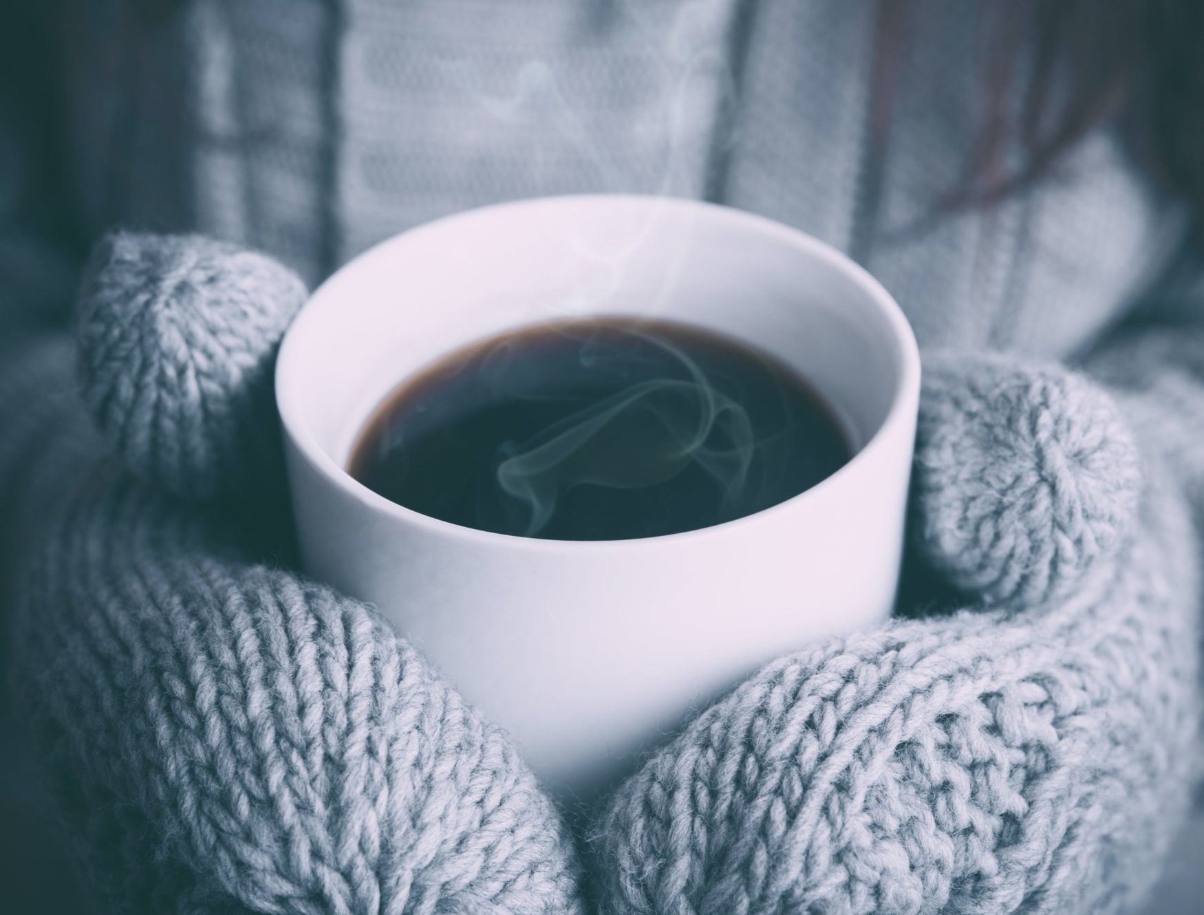 Two gray hands in gray mittens hold a white mug with a steamy dark liquid inside.