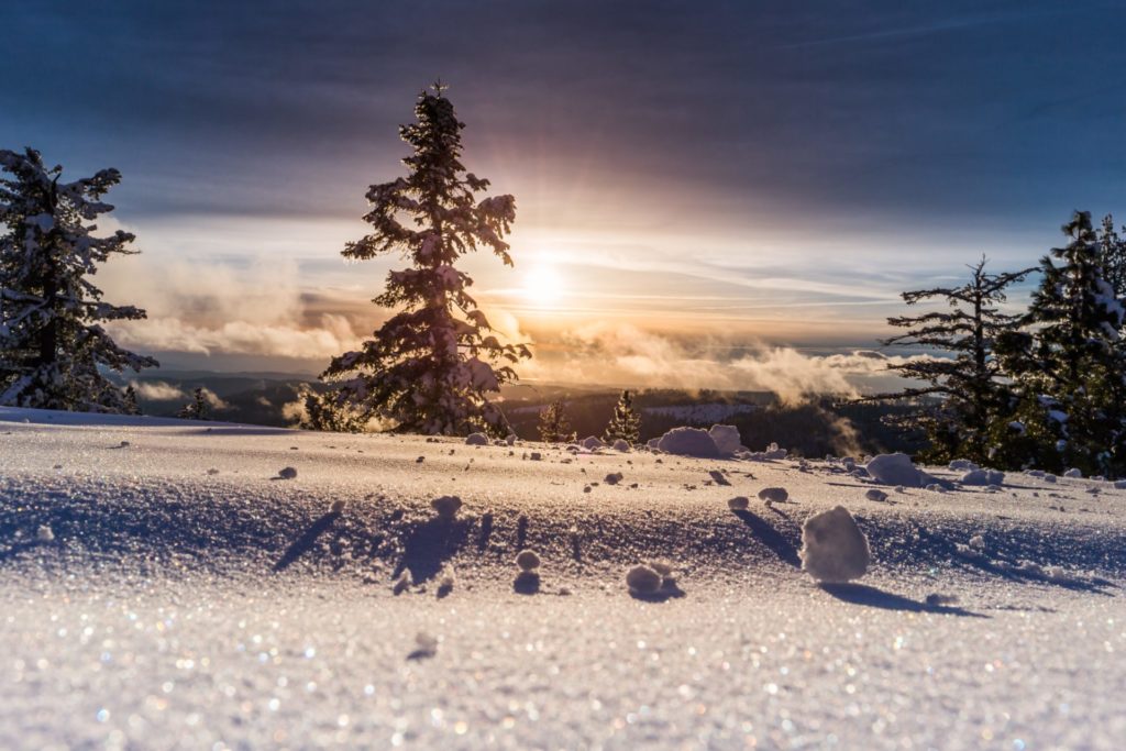 The sun can be seen over the clouds from a snowy mountain. Rolled snow casts shadows across the glittering landscape.