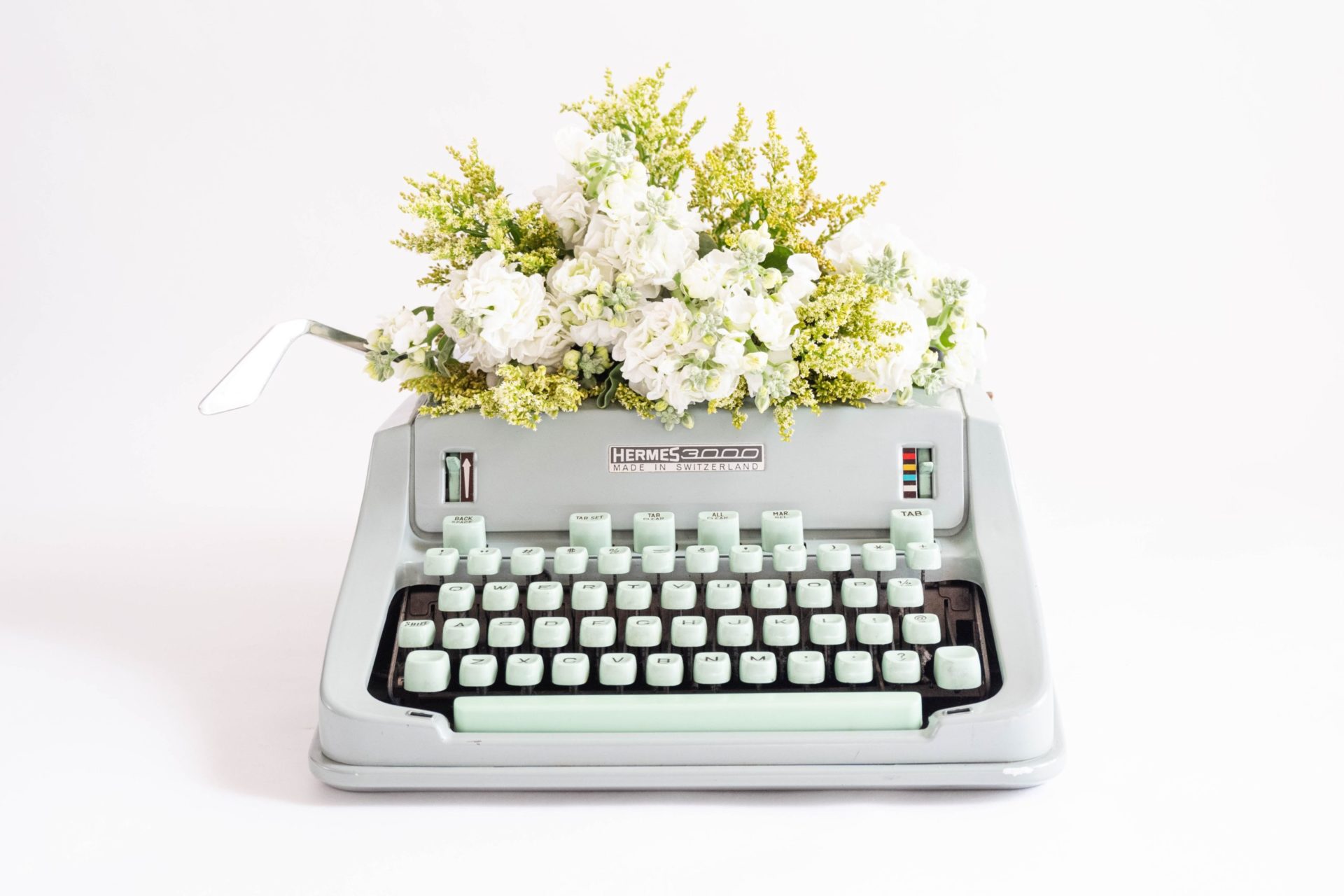 A light gray typewriter with pale blue keys sits on a white surface with white flowers and greenery poking out of the top.