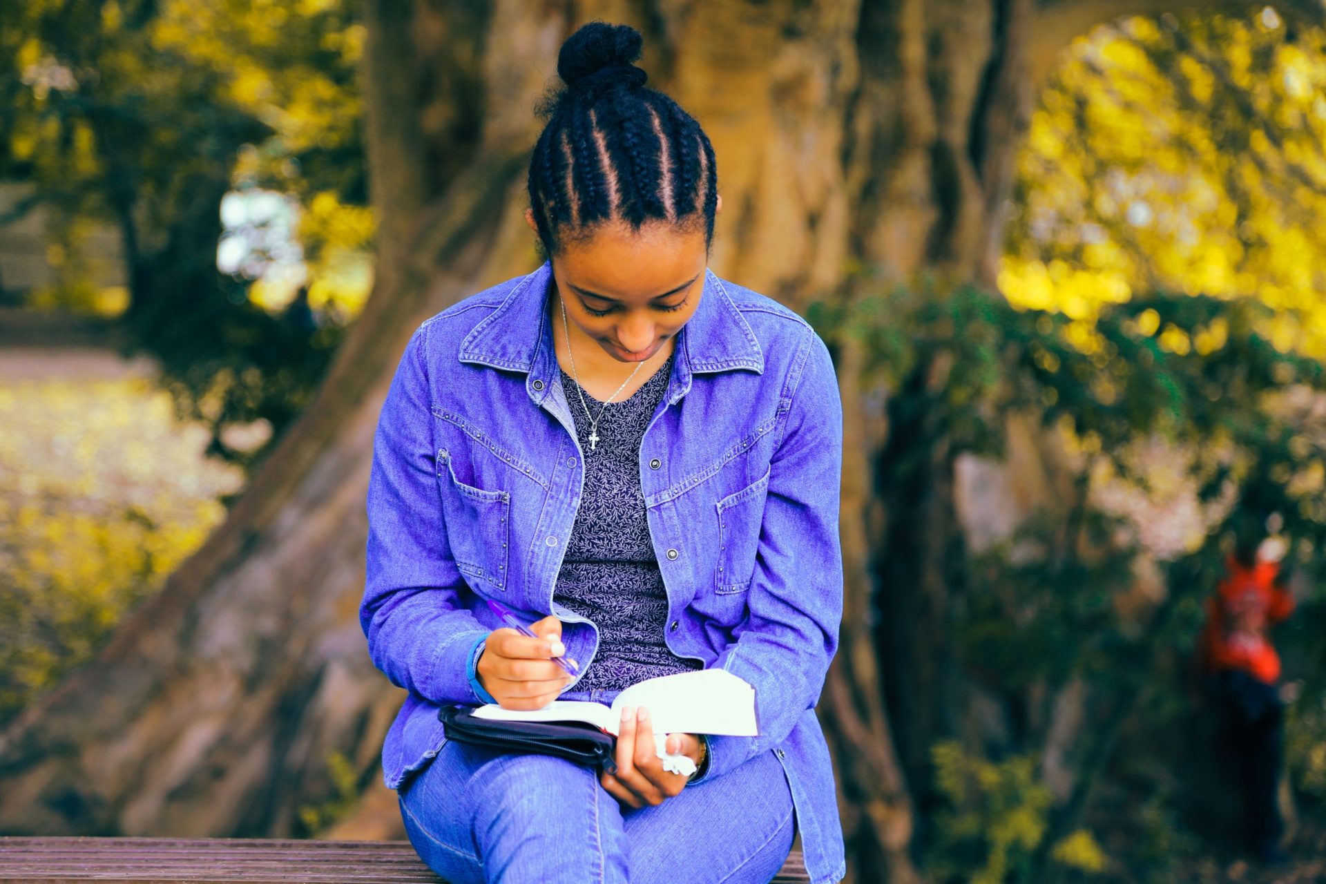 A young women with medium dark skin sits in front of a tree on a bench. She has braided hair that leads to a pony tail, a matching periwinkle jacket and pant and a gray-blue jacket. She is holding a purple pen with a book open on her lap.