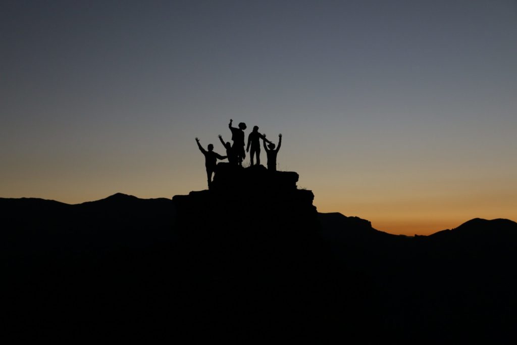 A silhouette of five people standing on a summit with an orange, yellow, and blue sky behind them. They are raising their hands and cheering.
