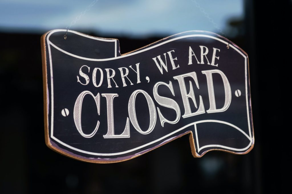 A black sign hang lettered with the words "Sorry, we are closed" hanging behind glass.