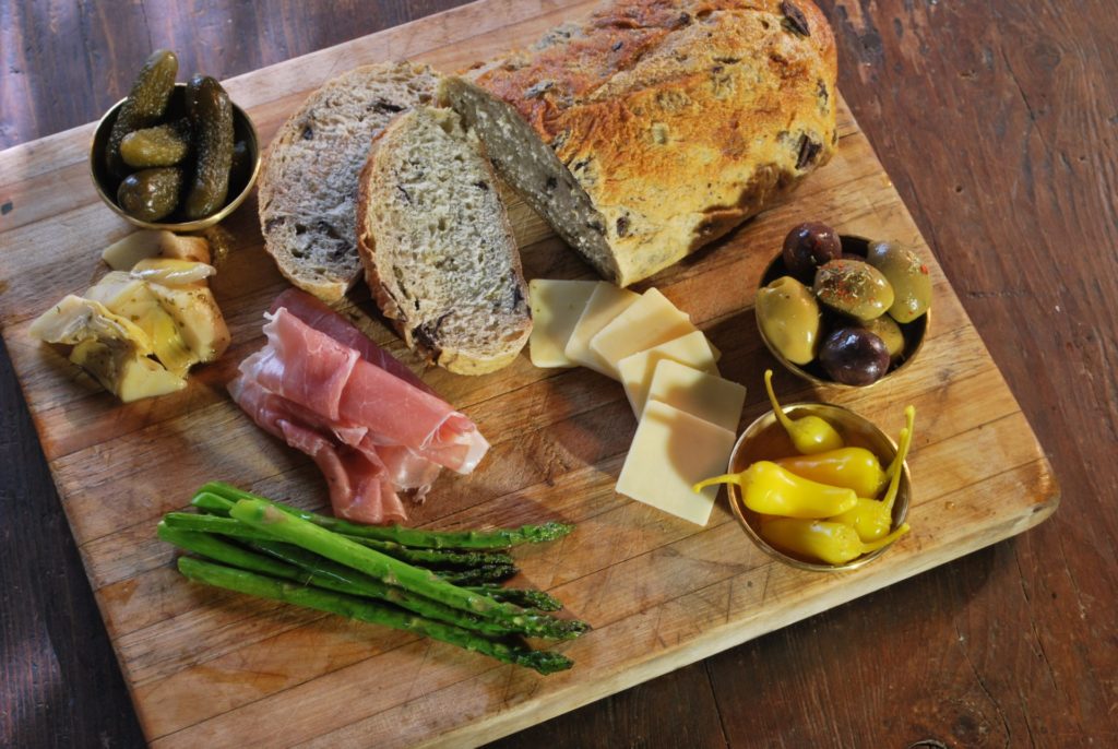 A wooden board with bread, meat, olives, cheese, and asparagus.