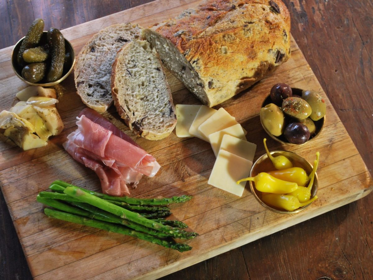 A wooden board with bread, meat, olives, cheese, and asparagus.