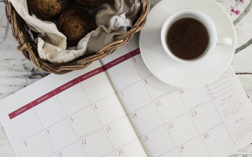 A dated planner open on a white tabletop with a cup of coffee in an espresso-sized cup in the corner. There is also a basket lined with a towel containing baked goods.